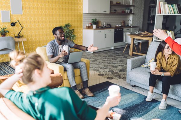 Cheerful multiethnic friends with drinks at home Smiling black young male with laptop sitting next to relaxing female friends with drinks watching crop faceless person at home